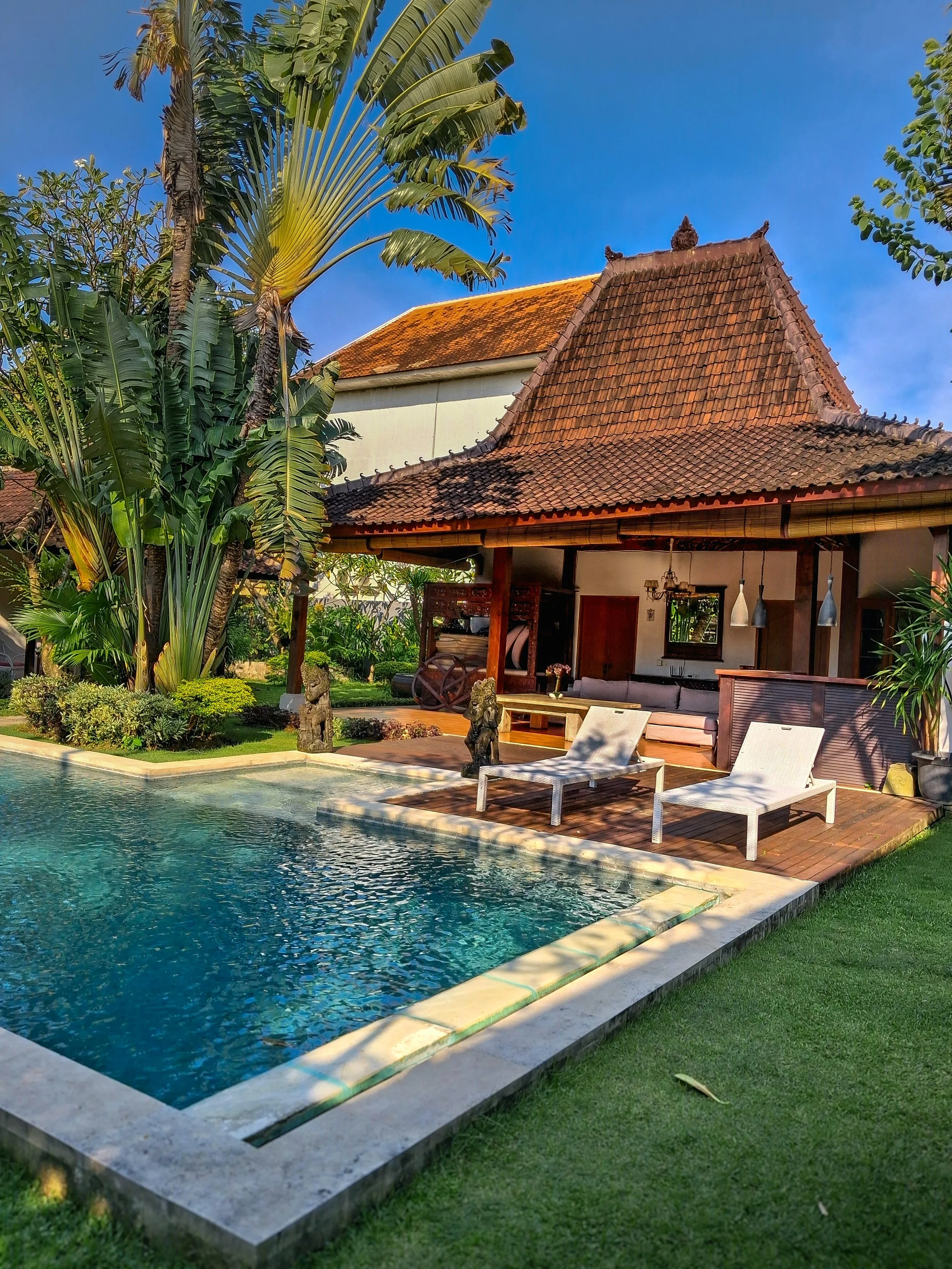 A backyard pool with clear blue water, surrounded by a concrete border and lush green grass. Two white lounge chairs are placed on a wooden deck by the pool. There is a traditional-style building with a steeply pitched roof covered in reddish-brown t