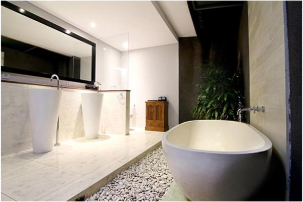Modern bathroom with a freestanding bathtub, two pedestal sinks with faucets, a large mirror, white pebbles along the edge of the tub, a small wooden cabinet, and lush green plants, with beige tiled flooring and a black wall.