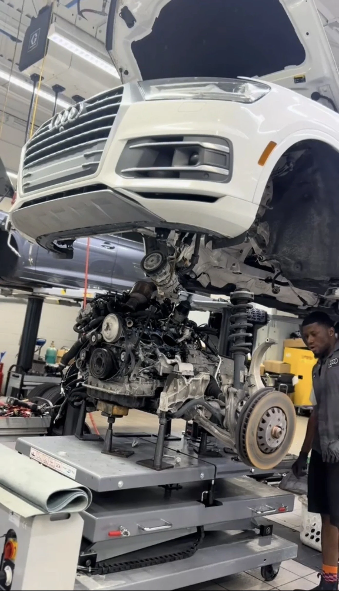 An automotive assembly plant with a white SUV on an elevated platform, being assembled or repaired with its front panels removed, exposing the engine and mechanical components. A technician is standing nearby.