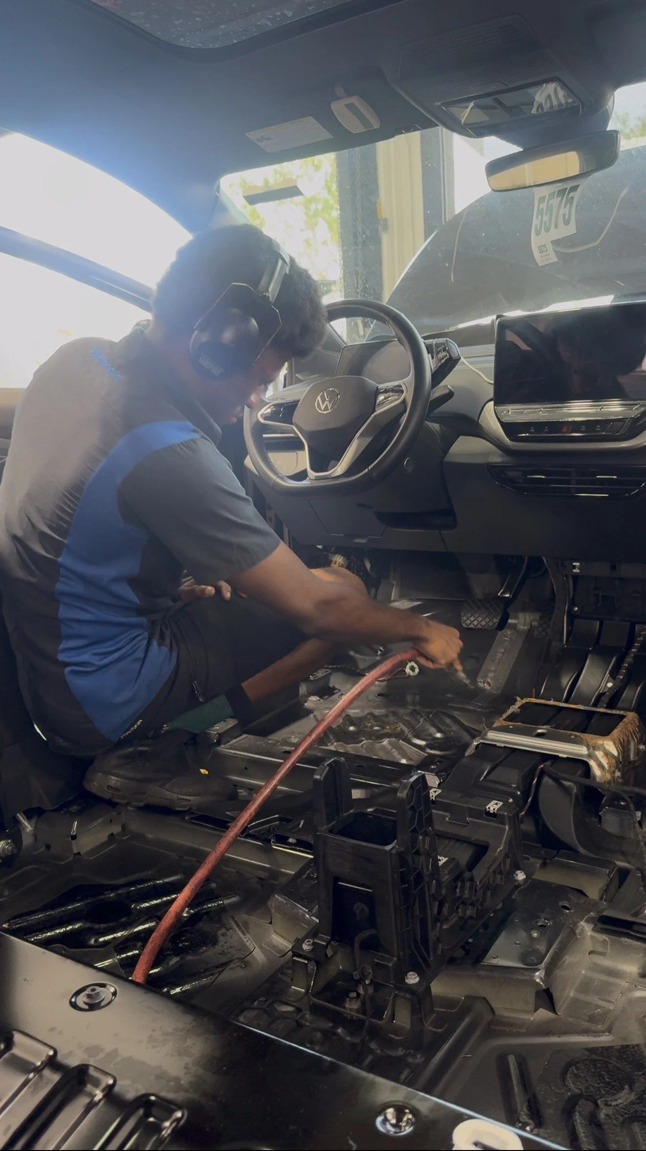A man working on the interior of a car, with the dashboard and floor disassembled, wearing a gray and blue shirt and large black headphones.