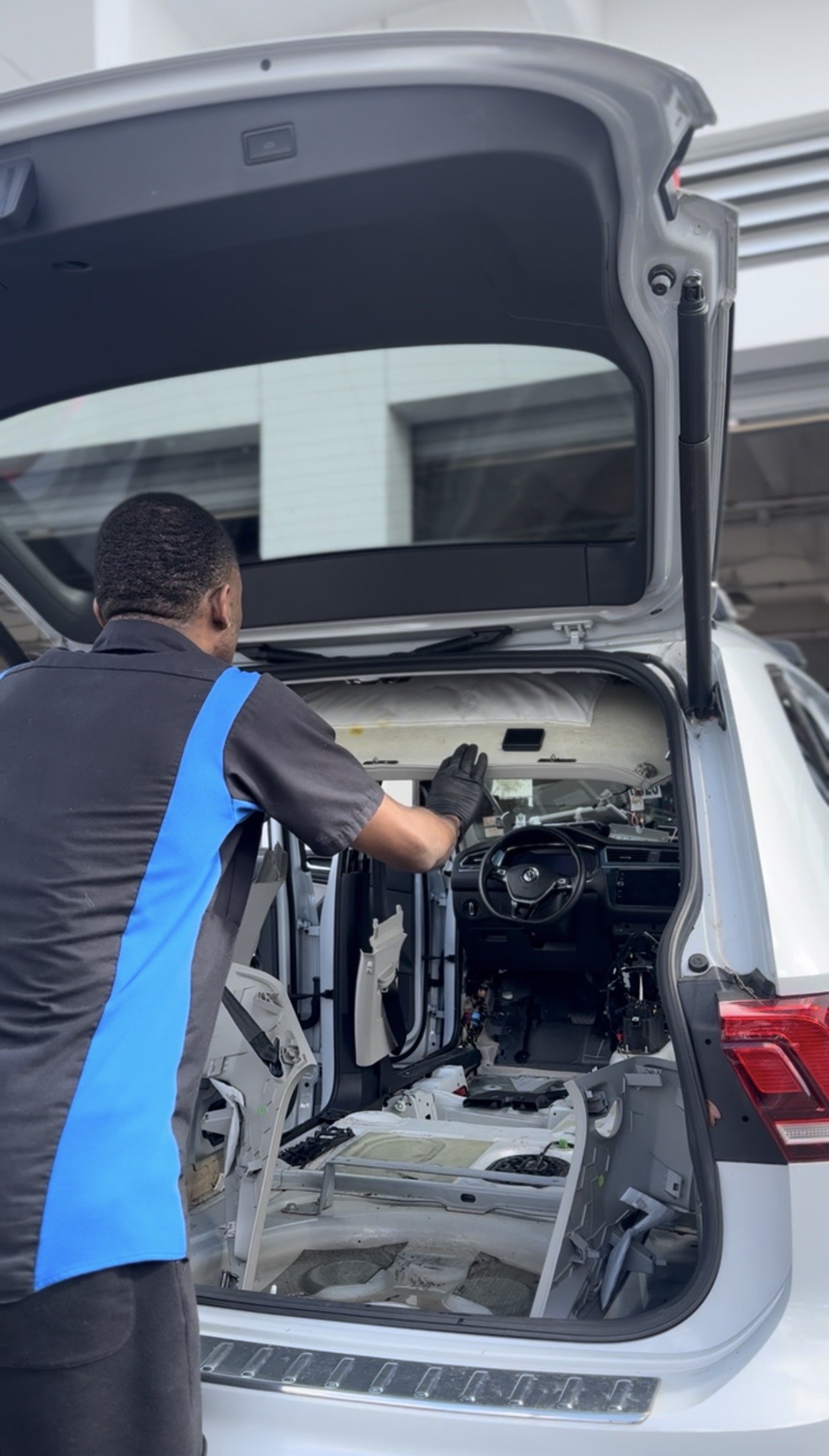 A mechanic or technician working on the interior of a vehicle with the rear hatch open, with parts and tools visible inside.