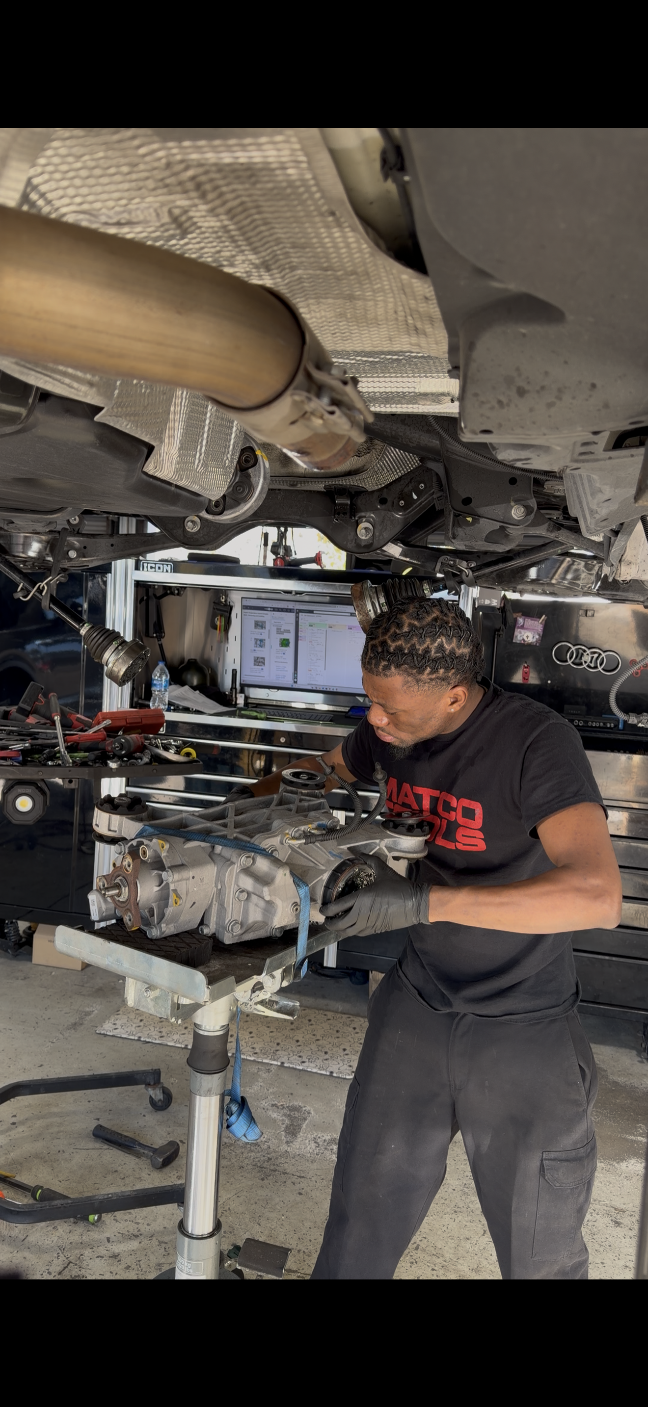 A mechanic working on a car engine in a garage, with tools and a computer monitor in the background.