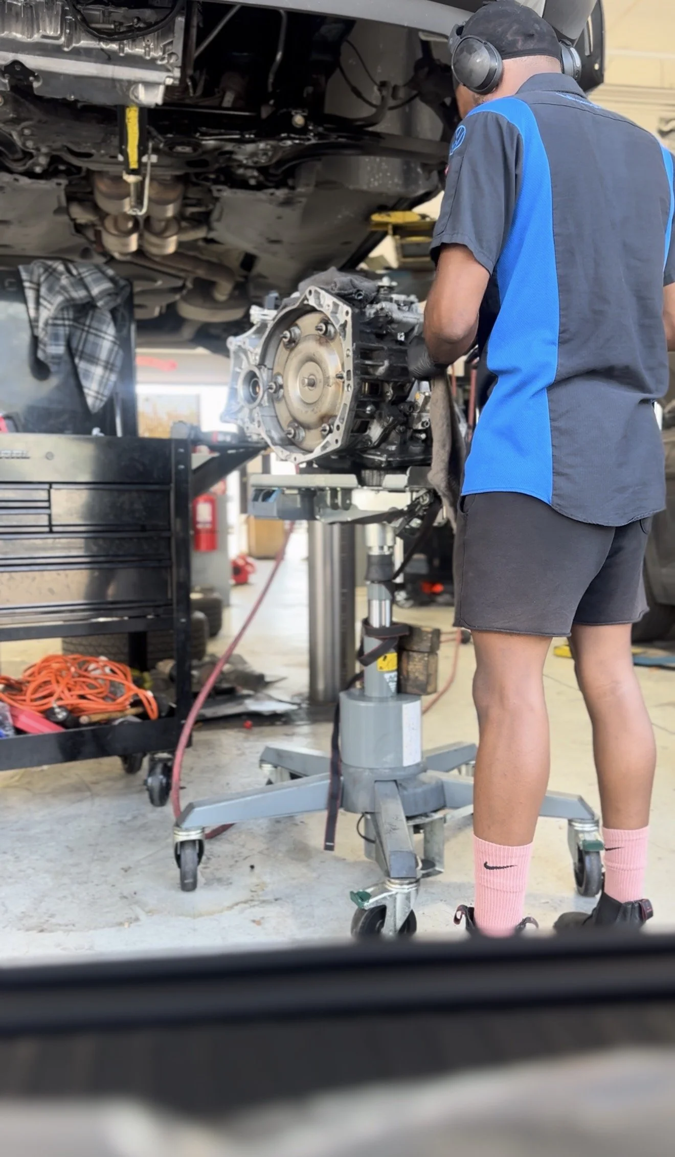 A mechanic working underneath a raised car in an auto repair shop, with an engine part on a stand and various tools and equipment nearby.