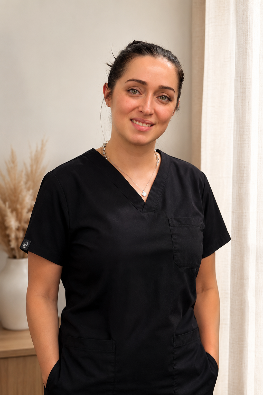 A woman wearing black medical scrubs with a necklace, standing indoors near cream-colored curtains and a vase with dried pampas grass.
