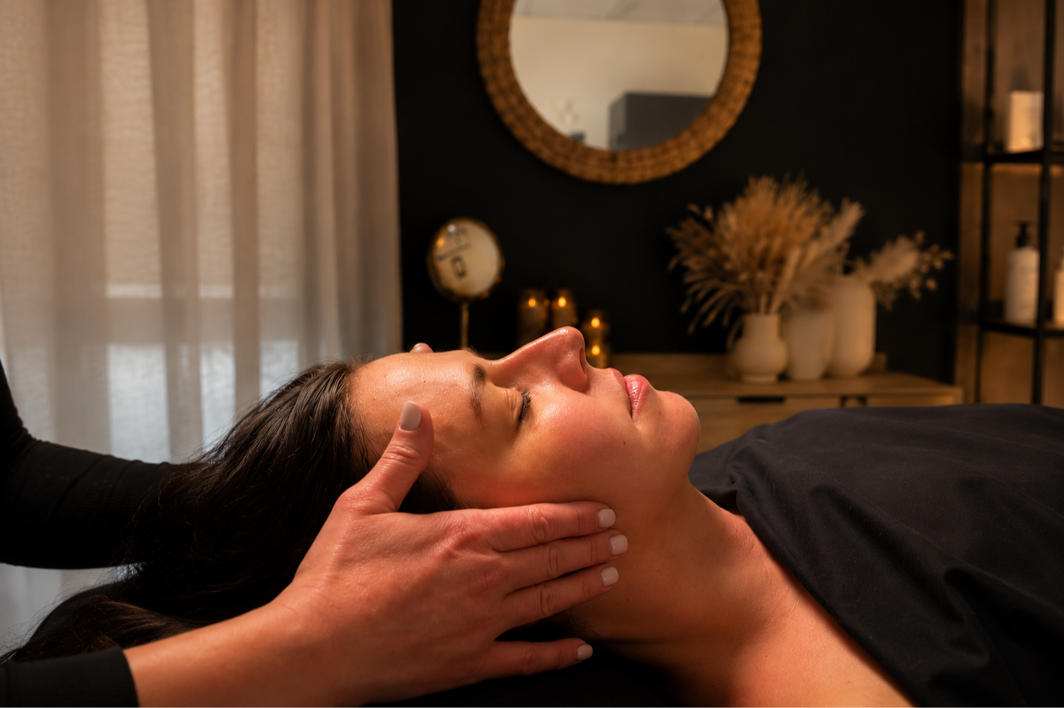 A woman receiving a facial massage in a spa room with a round mirror and decorative dried flowers in the background.