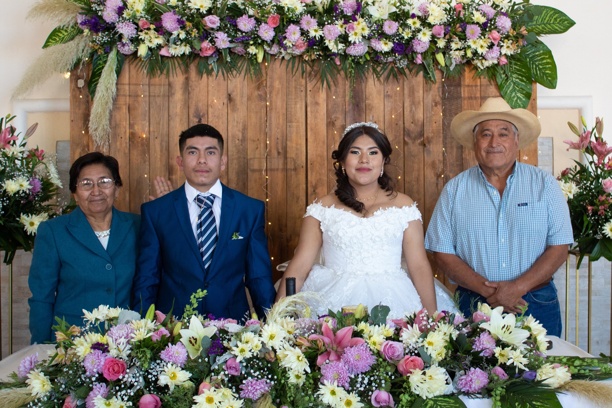 A group of five people, including a bride, groom, and three older adults, standing behind a floral arrangement at a wedding. The background features a wooden panel with purple, pink, white flowers, and greenery.