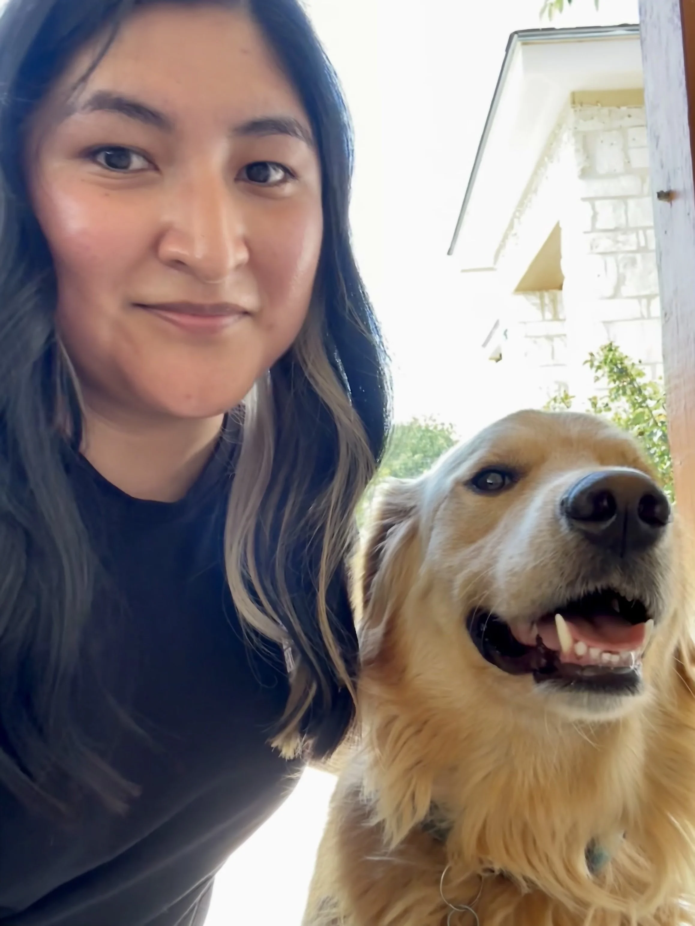 A woman with long dark hair and a dog with golden fur and a big smile, outdoors near a house with a brick wall and a window, on a bright, sunny day.