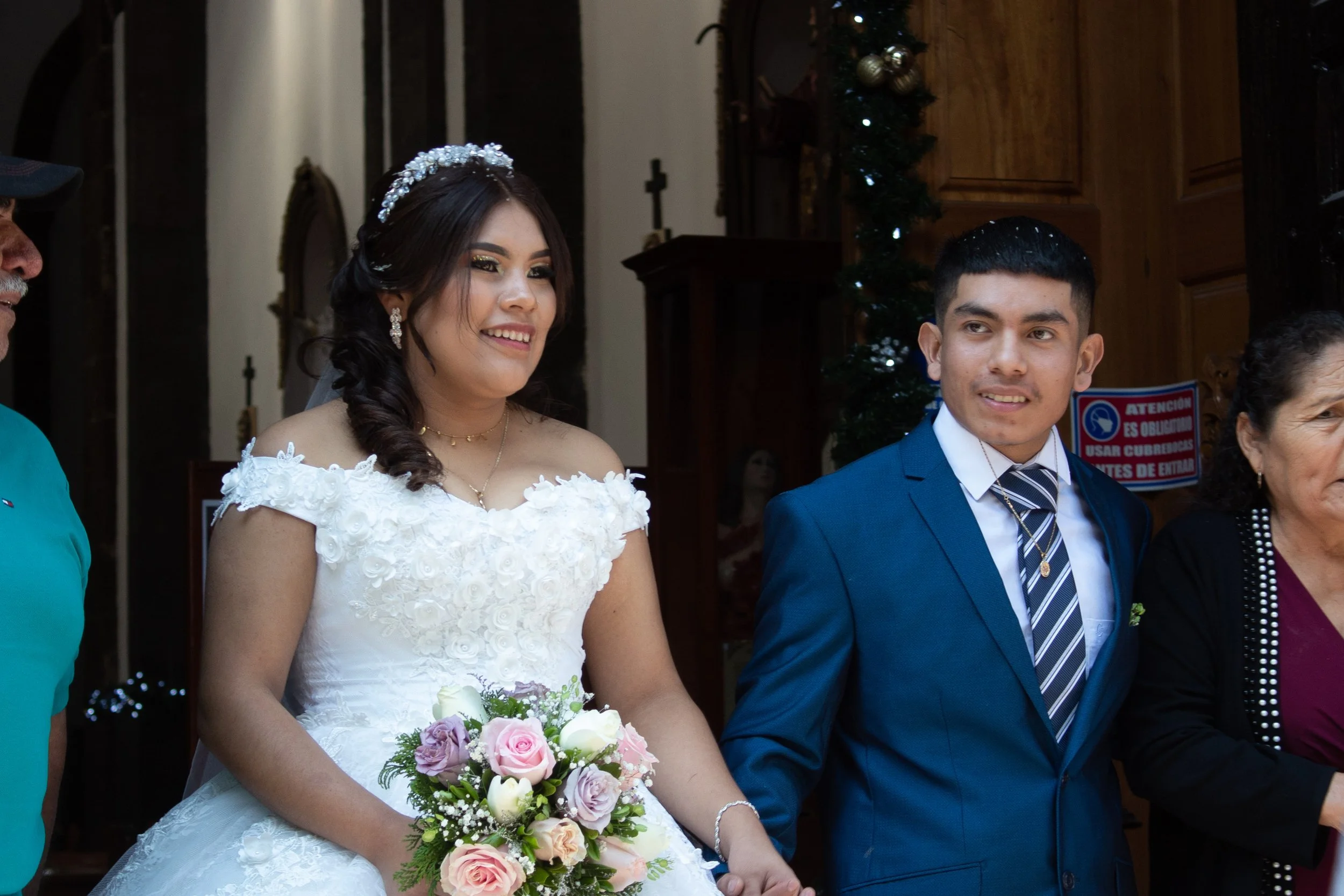 A bride in a white wedding dress holding a bouquet of pink and white roses and greenery, standing next to a groom in a blue suit during a wedding ceremony inside a church decorated with Christmas ornaments.
