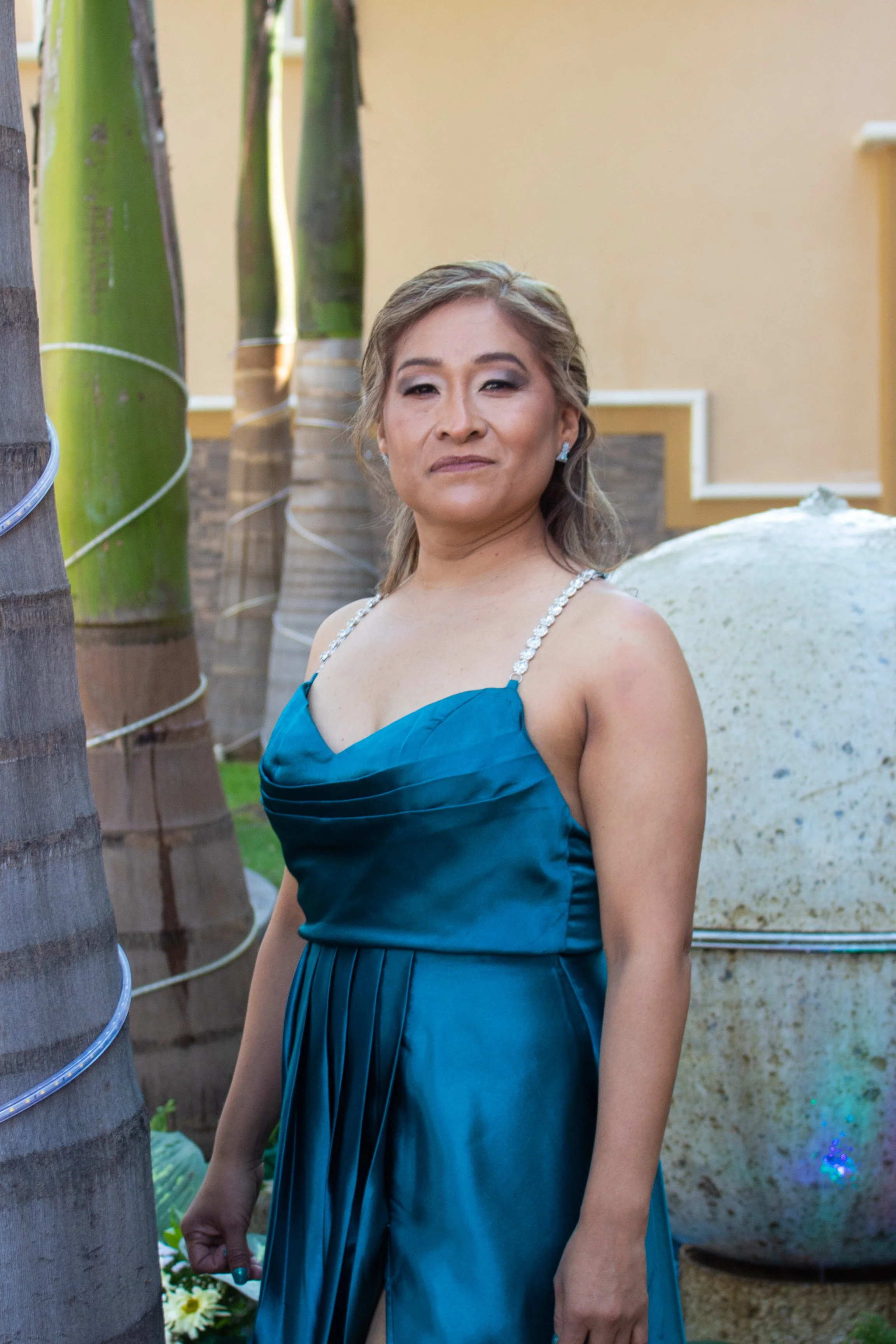 A woman in a blue strapless dress with gemstone straps standing outdoors near bamboo and a large vessel, with a beige wall in the background.