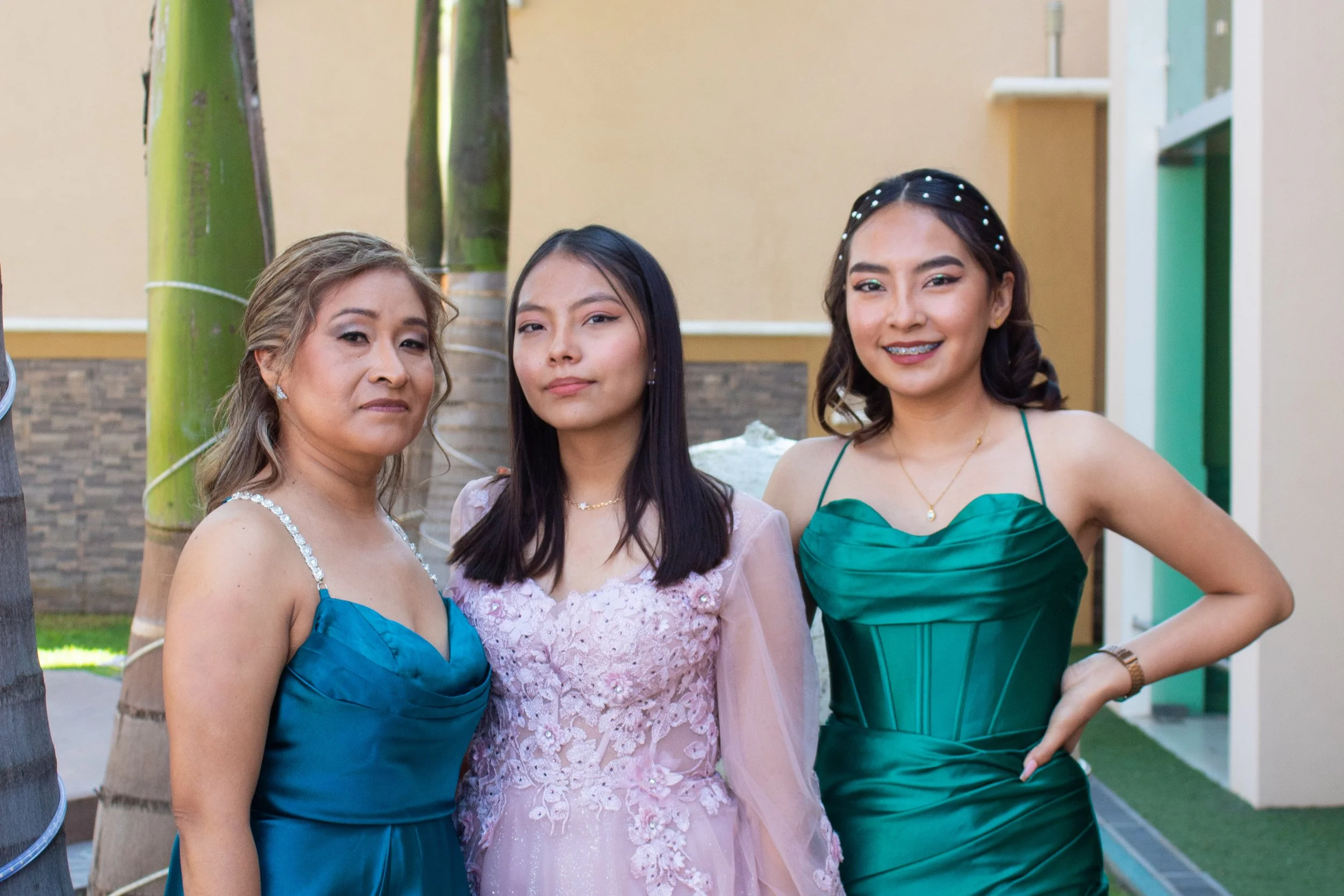 Three women standing outdoors at a formal event, wearing elegant dresses. The woman on the left has blonde hair styled in loose waves and is wearing a blue dress with a beaded strap. The woman in the middle has dark straight hair and is dressed in a light pink lace gown. The woman on the right has dark curly hair with pearl accents and is wearing a green satin dress with thin straps, smiling at the camera.