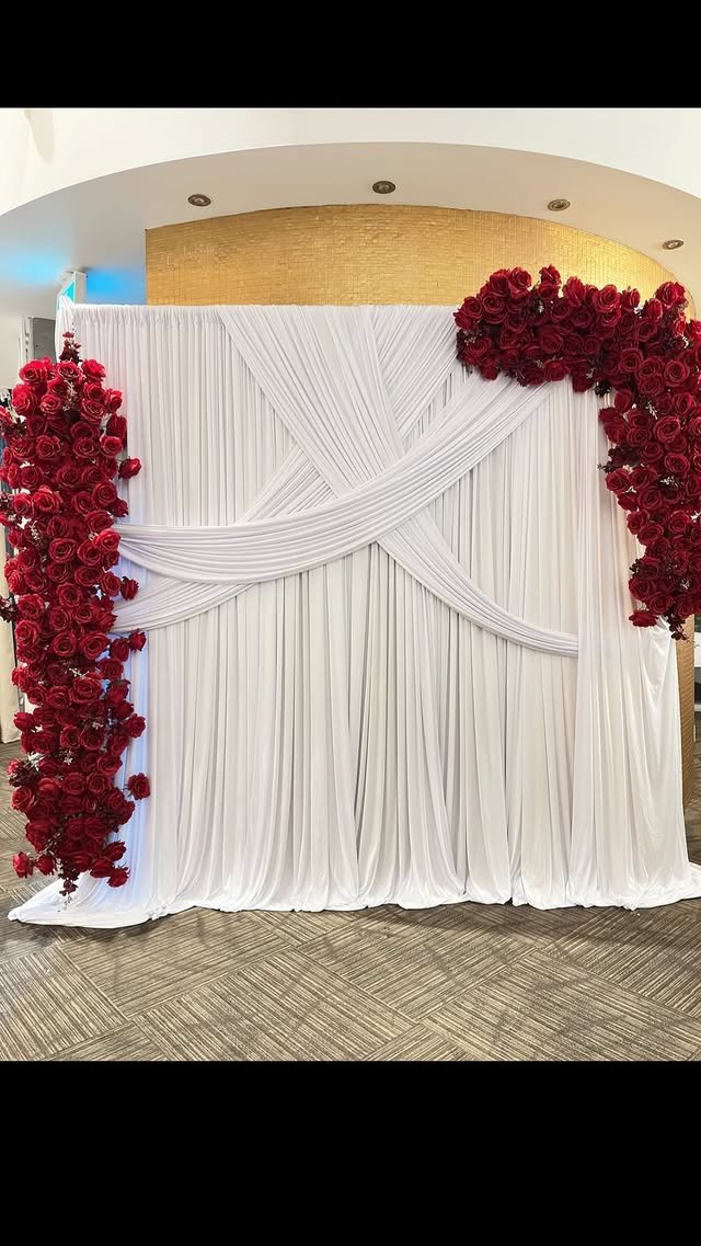 A white fabric backdrop adorned with red roses arranged along the sides and top, with pleated drapery and flowing fabric details for a wedding or event setting.