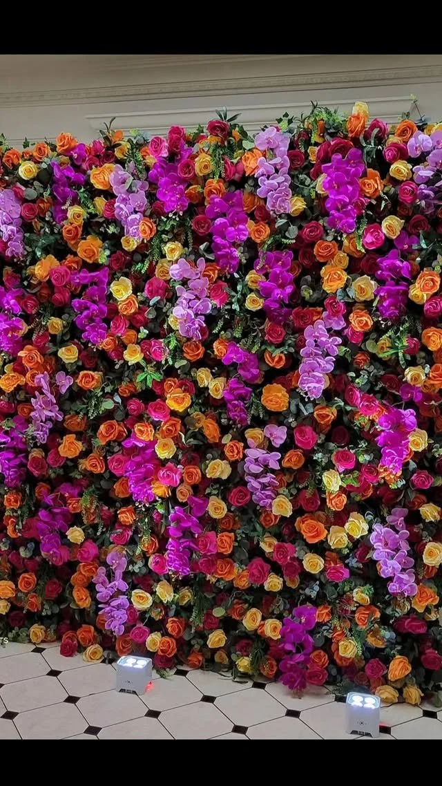A vibrant wall of artificial roses and orchids in orange, pink, yellow, and purple, with artificial greenery behind, situated on a black and white tile floor with two small white spotlights in front.
