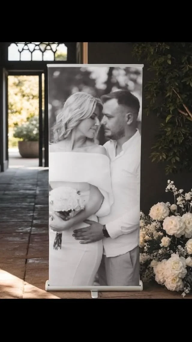 Black and white photograph of a couple on their wedding day, with the bride holding a bouquet and the groom embracing her, displayed on a standing poster.