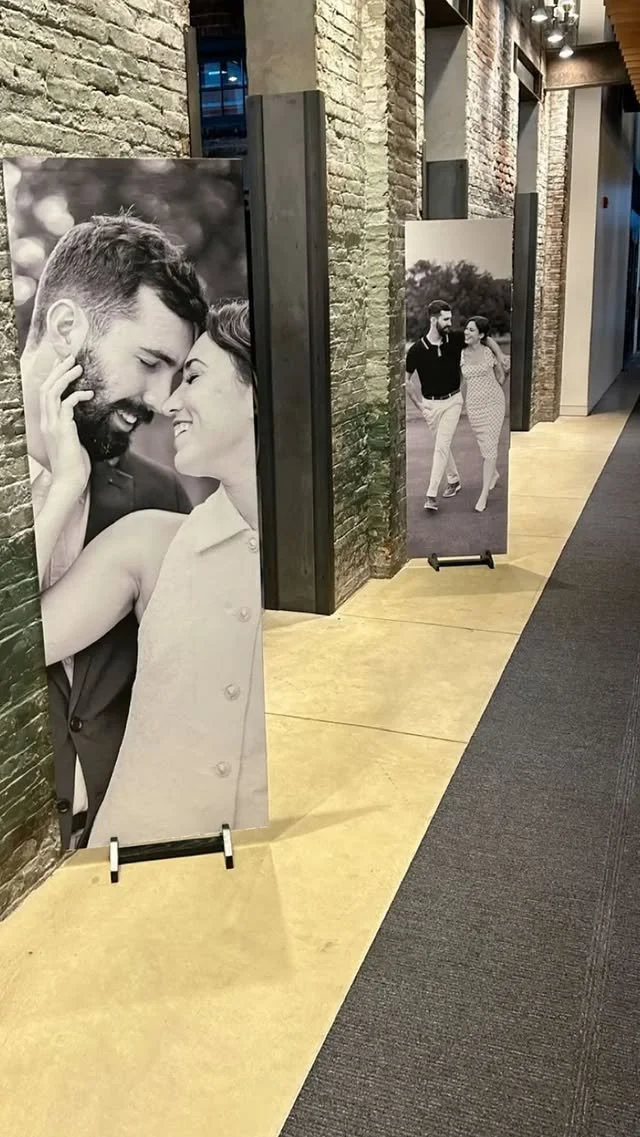 Black and white photos of couples on display in an indoor gallery or corridor, with exposed brick walls and framed images.