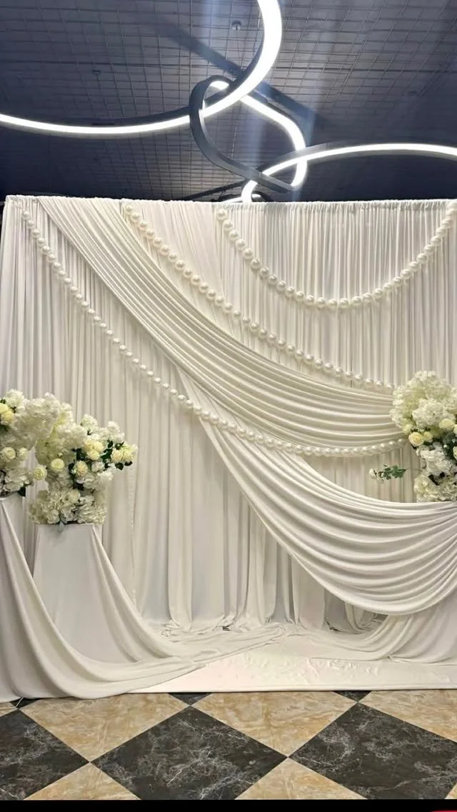 Elegant wedding backdrop with white draped fabric, floral arrangements with white flowers, and decorative light fixtures overhead.