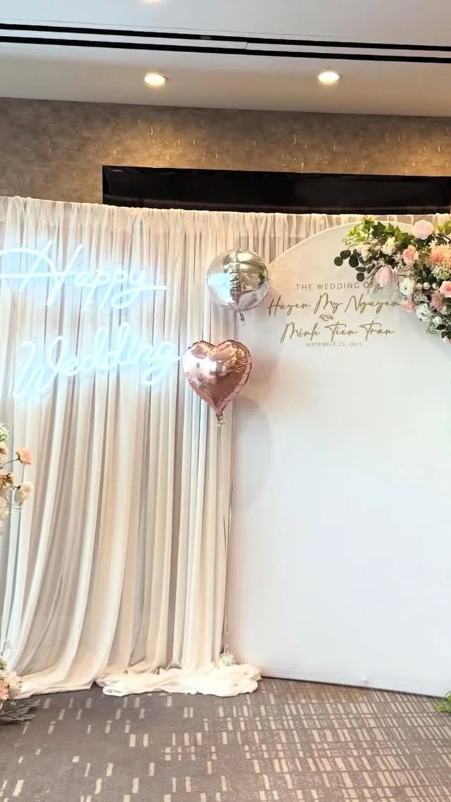 Wedding backdrop with white drapes, pink and silver balloons, and a floral arrangement, with a neon sign saying 'Happy Wedding'.