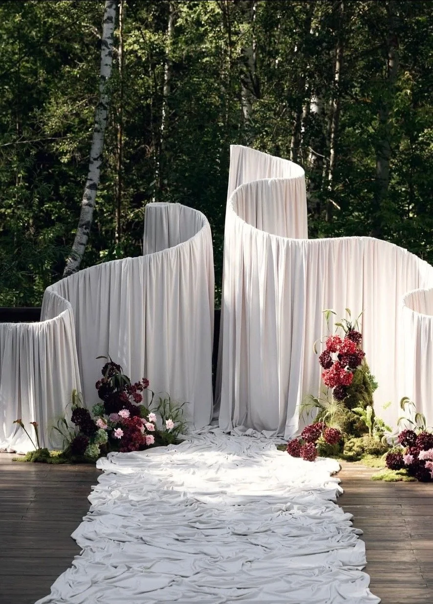 White draped fabric panels creating a semi-circle backdrop in a forest setting, with a white fabric aisle and pink and purple flowers decorating the sides, likely for a wedding ceremony.