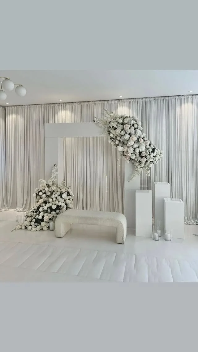 A white wedding altar adorned with large floral arrangements of white flowers and greenery. The backdrop features white curtains, with a white bench at the center and clear cylindrical vases on stands to the right.