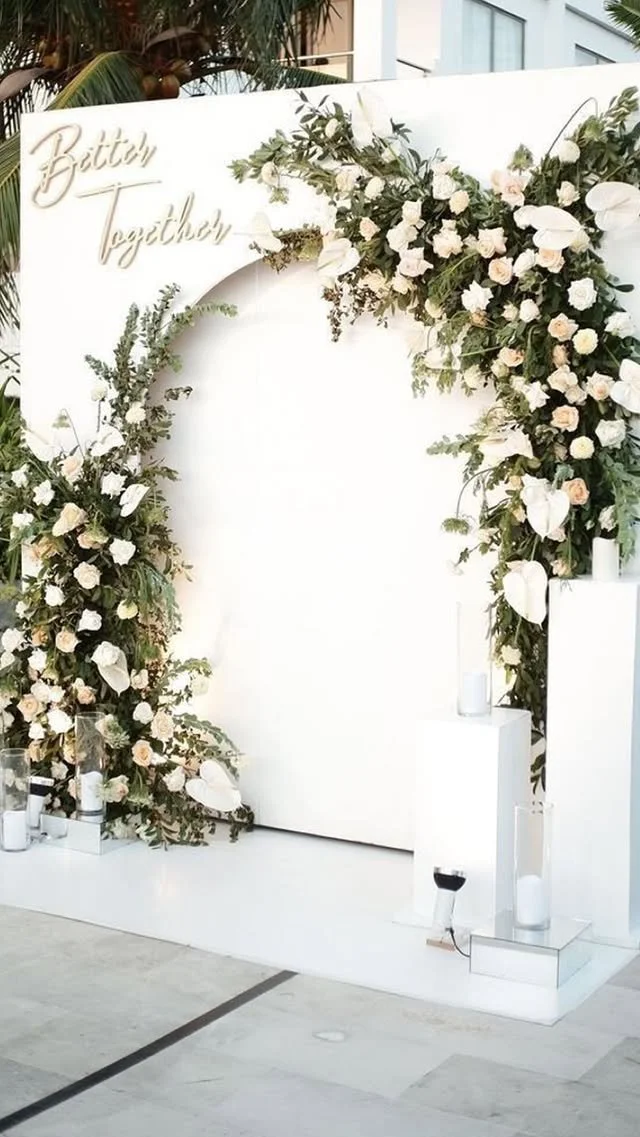A floral wedding arch decorated with white and soft pink roses, green foliage, and white calla lilies, with the words "Bitty Together" in cursive at the top, set on a white background with candles in glass holders at the base.