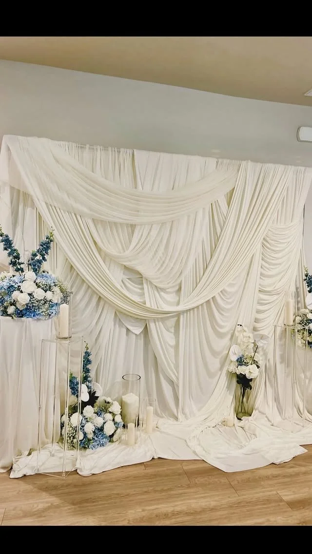 Elegant wedding backdrop with white draped fabric, floral arrangements in blue and white, and candles.