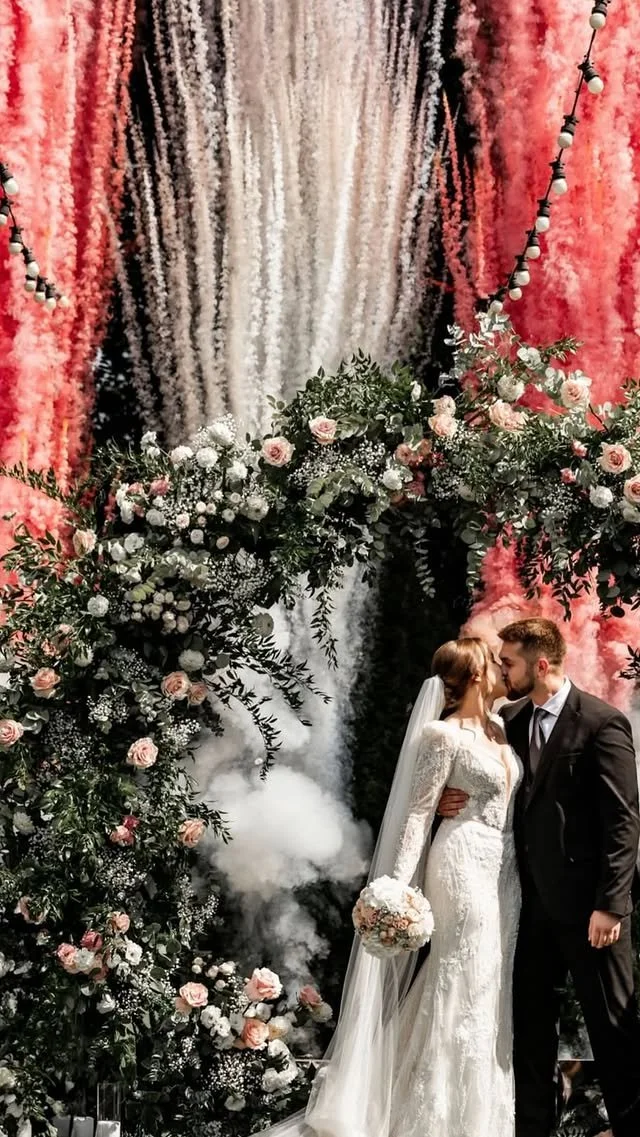 A bride and groom share a kiss during their wedding ceremony, surrounded by an elaborate floral arch with pink and white flowers and hanging decorations.