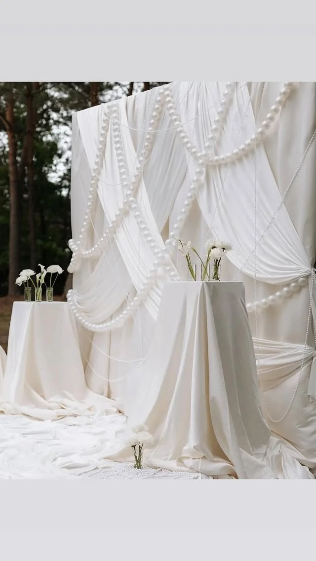 Elegant outdoor event setup with white draped fabric, pearl garlands, and small tables with white flowers in glass vases.