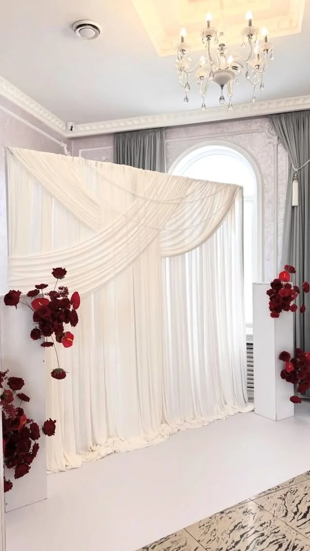 Elegant indoor event space with a white draped backdrop, red floral decorations, a chandelier, and large window with gray curtains.