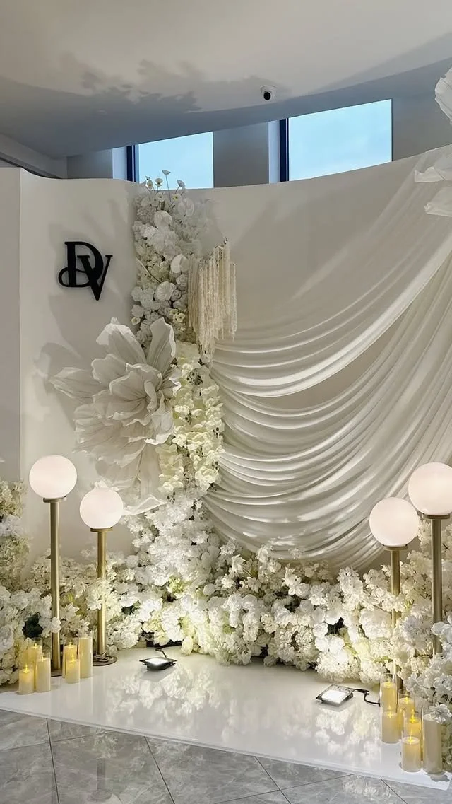 Elegant white floral backdrop with draped fabric, surrounded by white flowers, gold and white lamps, candles, and a miniature black dish, likely for a wedding or special event.