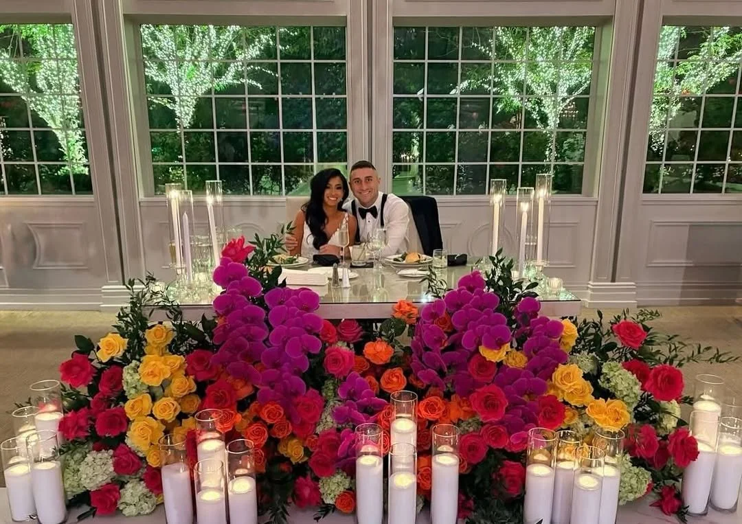 A newlywed couple sitting at a decorated reception table with pink, yellow, and orange flowers and white candles, in front of large windows with string lights on trees outside.