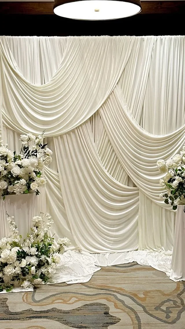 Elegant wedding backdrop with cream-colored draped fabric and floral arrangements with white roses and greenery.