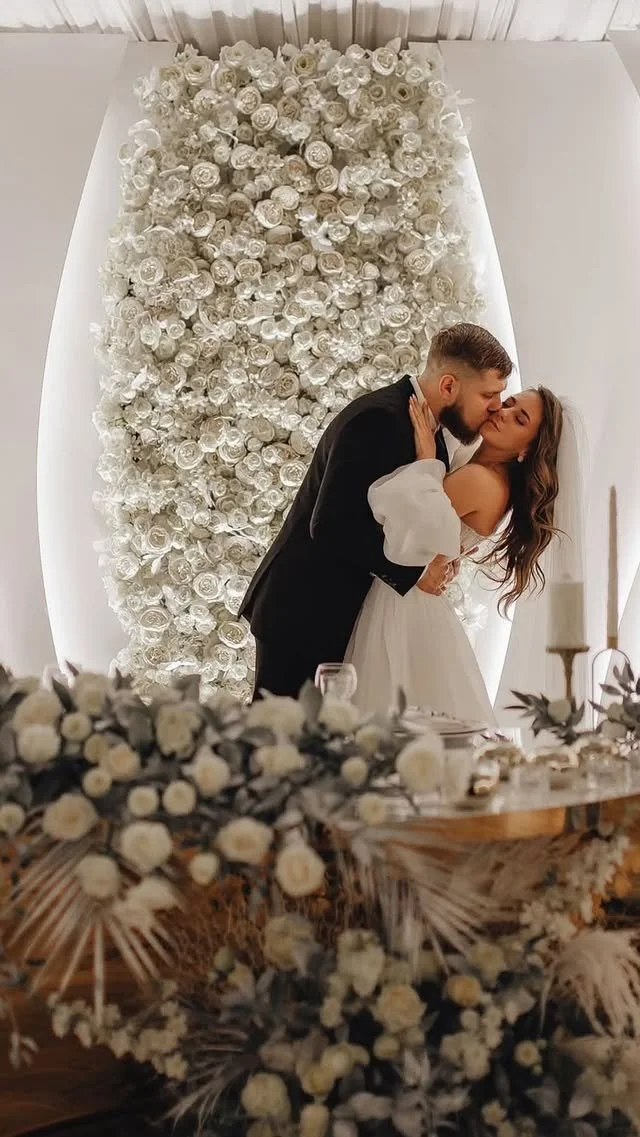 A bride and groom sharing a kiss during their wedding ceremony, with a backdrop of white roses and flowers.