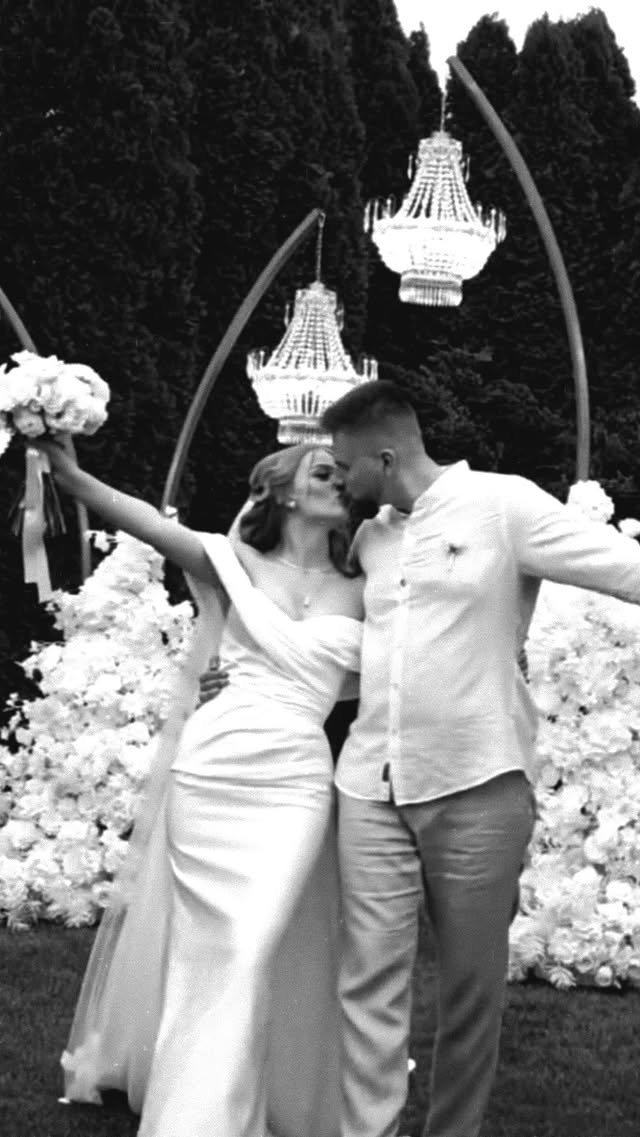 A couple is kissing at a wedding or romantic event, with the woman wearing a wedding dress and holding a bouquet, while the man is dressed in a casual shirt and pants. They are standing outdoors with large flowers and two ornate chandeliers hanging i