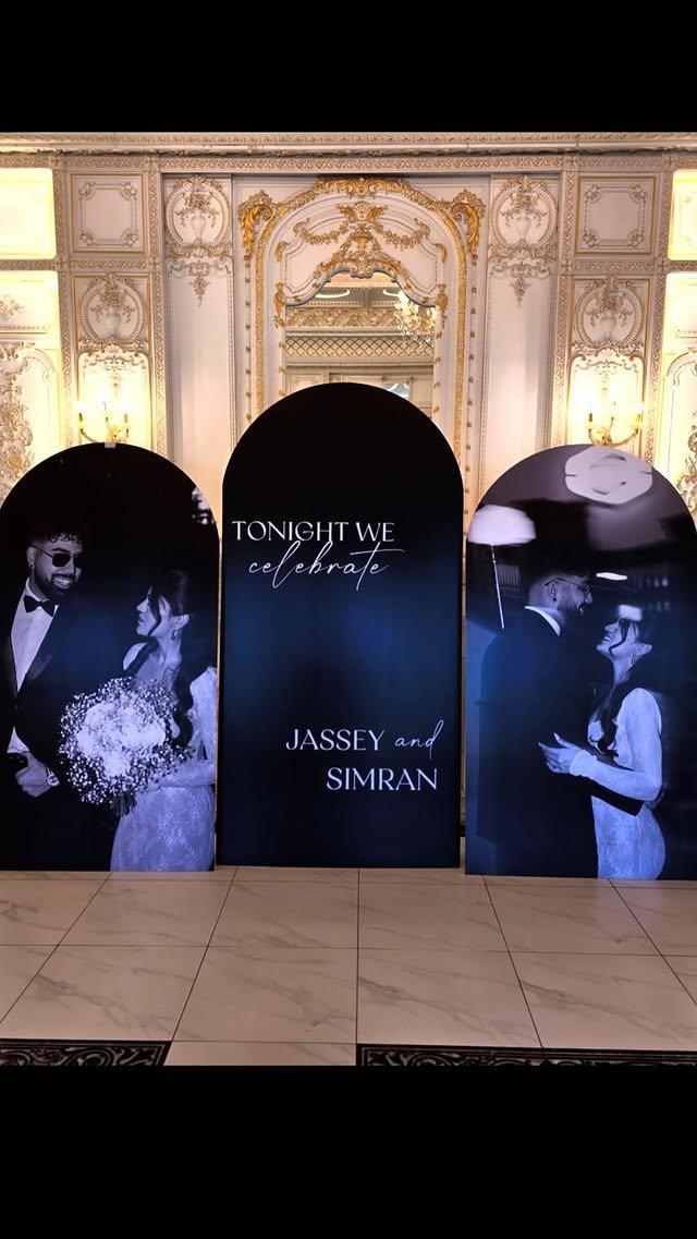 Decorative wedding display with black arches featuring black-and-white photos of a bride and groom, central arch with text 'Tonight We Celebrate Jassey and Simran', ornate white and gold interior background.