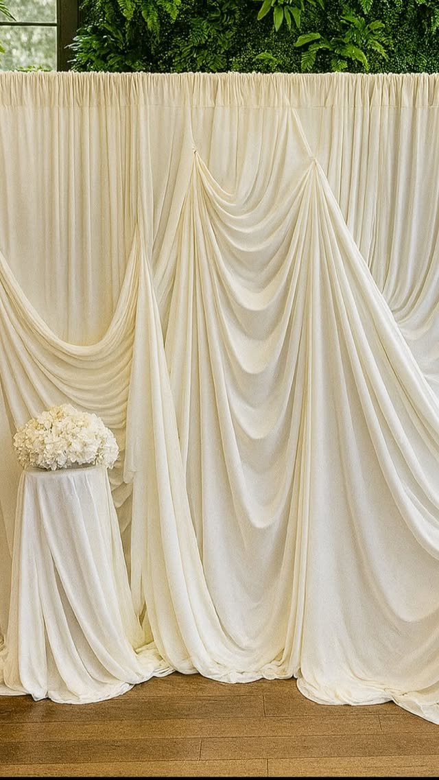 Cream-colored draped fabric backdrop with a small table holding a bouquet of white flowers in front of a window and green foliage.