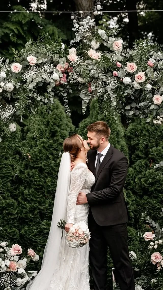 Bride and groom sharing a kiss during their wedding ceremony, with floral decorations and greenery in the background.