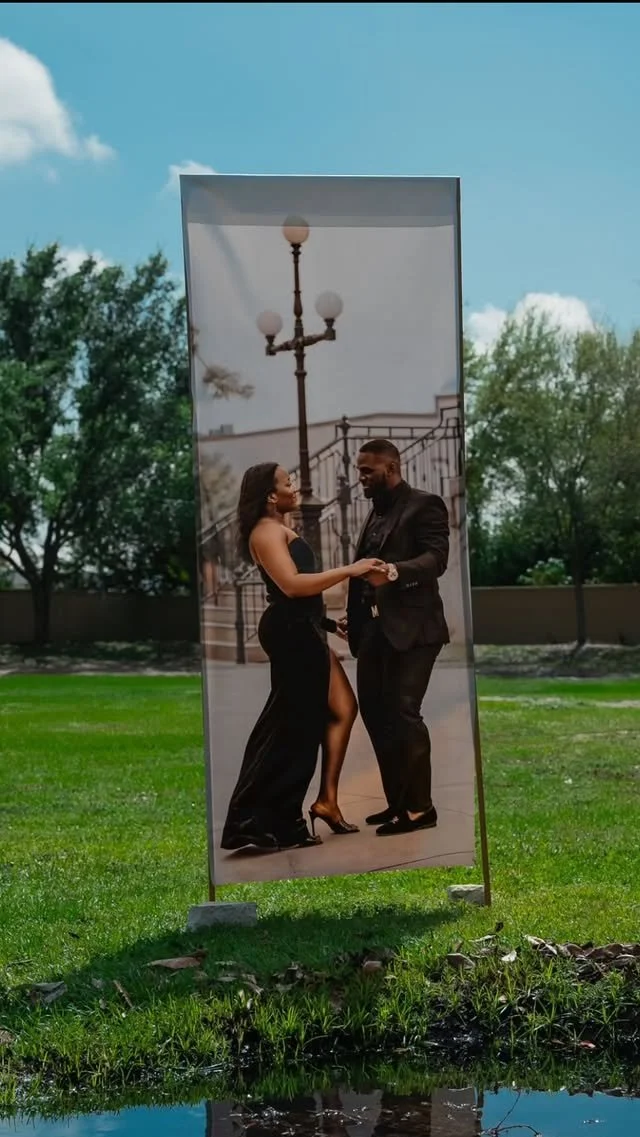 A portrait of a man and woman dancing outdoors in front of a large photo display, with trees and blue sky in the background.