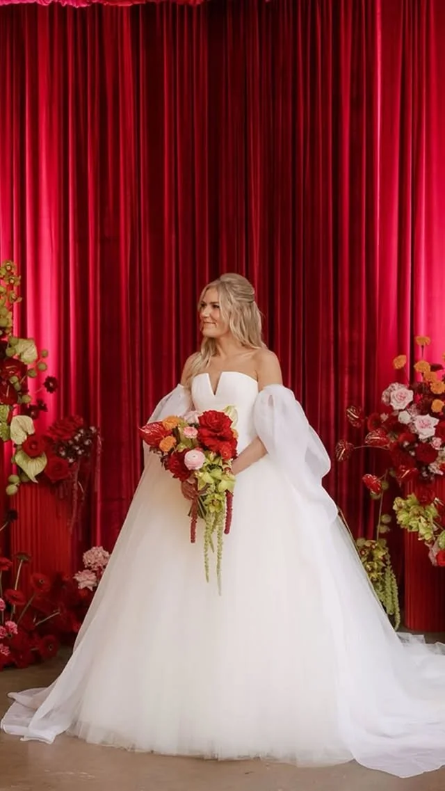 A woman in a white wedding dress holding a bouquet stands in front of a red curtain with flower arrangements on each side.