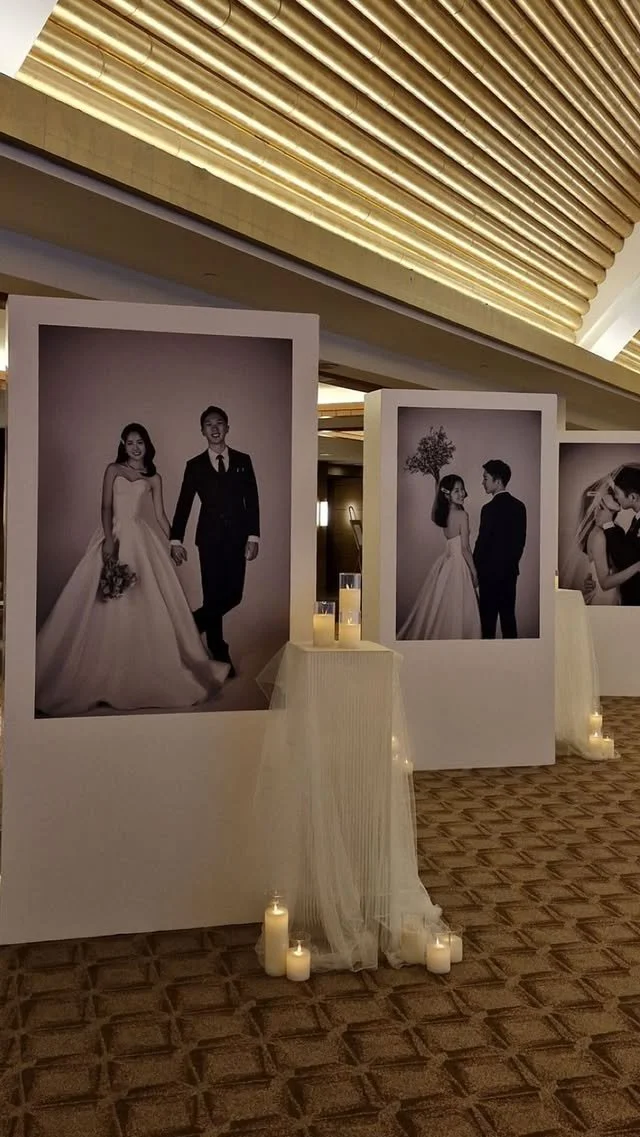 Exhibition of large black-and-white wedding photographs displayed on white panels, with candles on the floor and table, in an elegant interior setting.