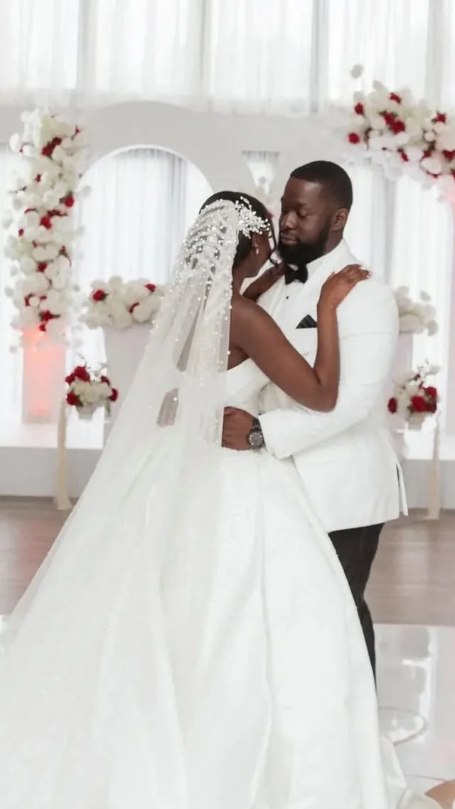 A bride and groom share a dance at their wedding in a decorated reception hall with white and red floral arrangements.