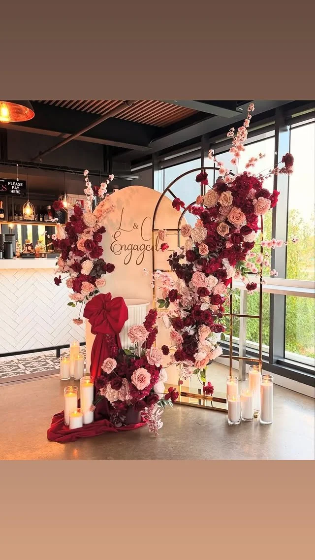 Floral wedding backdrop with pink, red, and white flowers, candles, and a sign that reads "L & Engage" in a modern indoor setting with large windows.