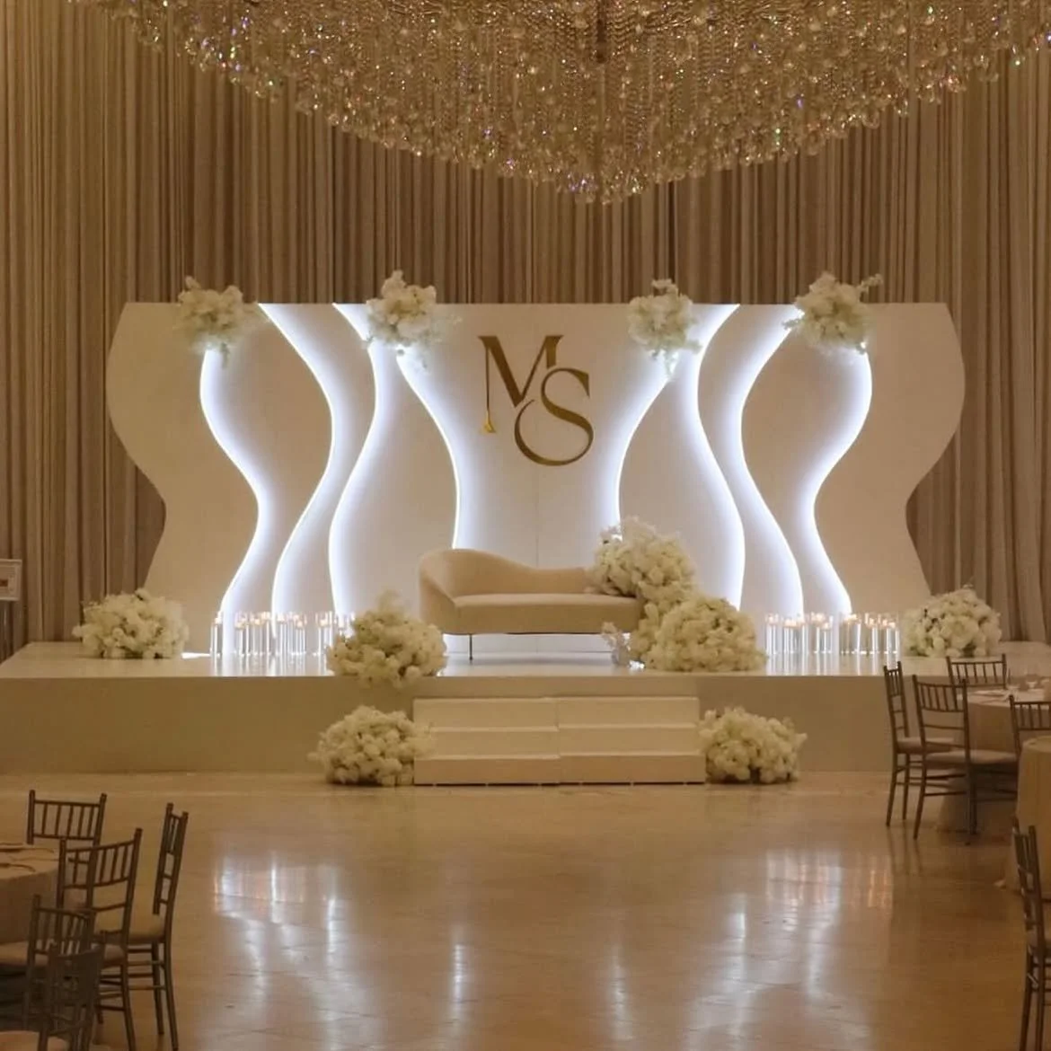 Elegant wedding stage with cream-colored backdrop, illuminated with soft white lighting, featuring white floral arrangements and a central white chaise lounge, with overall decor in warm, neutral tones.