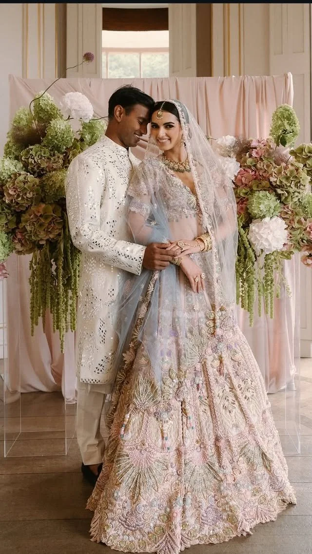A bride and groom in traditional wedding attire standing together, smiling, in front of a floral backdrop at their wedding.
