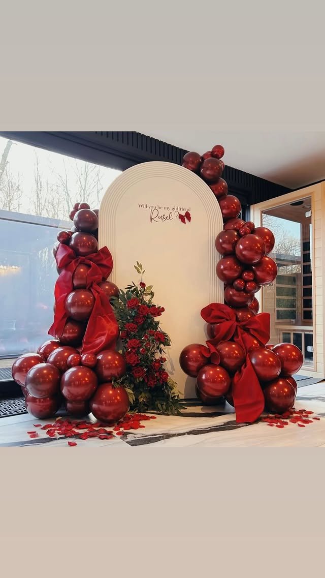 Valentine's Day themed backdrop with red balloons, red bows, rose petals, and a floral arrangement, with a sign that asks 'Will you be my girlfriend Rusel?'