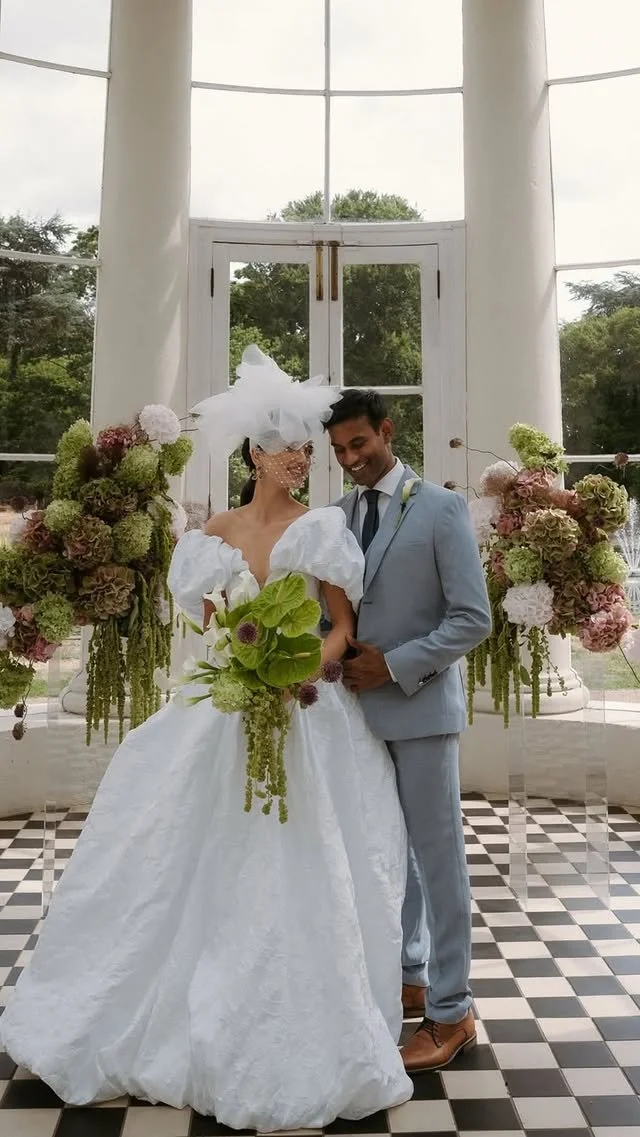 A bride and groom standing inside a bright, white room with large windows, decorated with floral arrangements, sharing a joyful moment on their wedding day.