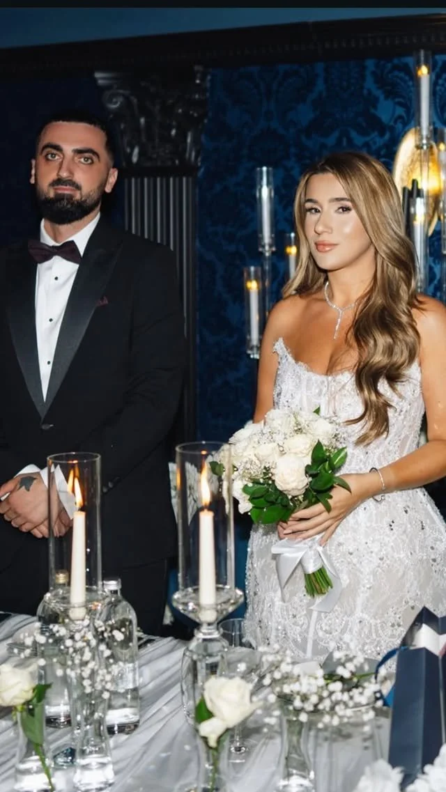 A bride in a white lace wedding dress holding a bouquet of white roses, standing next to a groom in a black tuxedo with a bow tie, at a wedding reception with candles and floral decorations.