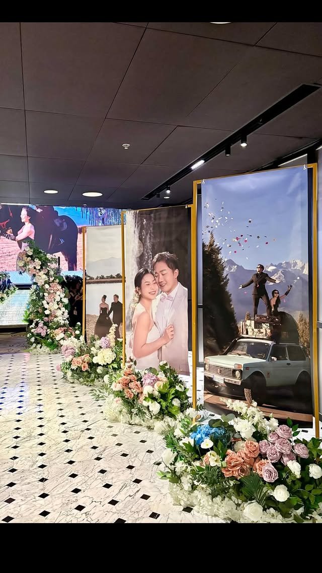 Display of large photos and floral arrangements at a wedding or event venue, including floral decorations on the floor, with a couple in wedding attire in one image and an outdoor scene with a car and people on top in another.