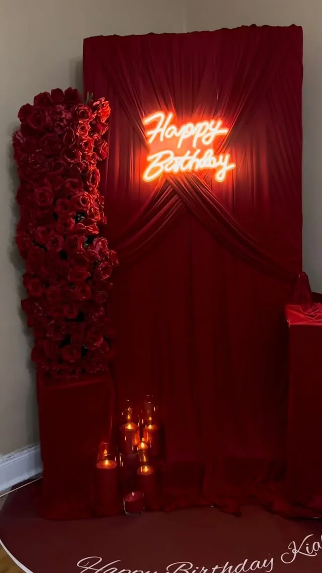 Red floral birthday backdrop with a neon sign that says 'Happy Birthday,' red drapery, glowing candles on the floor, and a small red table on the right.