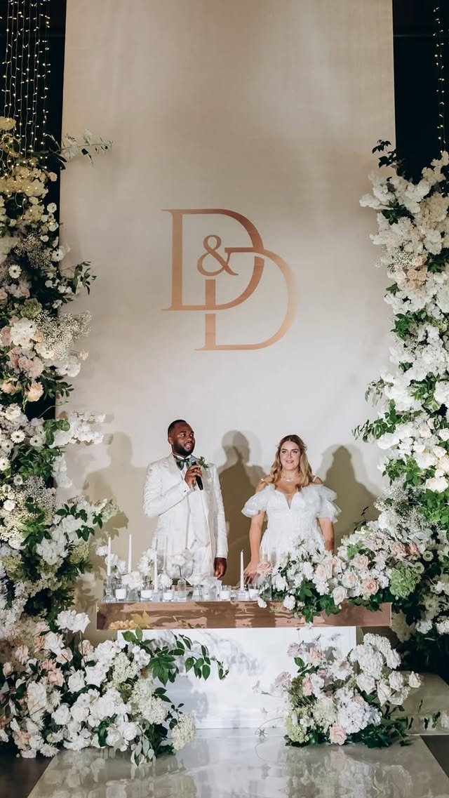 A wedding reception with a bride and groom standing behind a decorated table. The groom is speaking into a microphone while the bride looks on. The backdrop features large floral arrangements and a monogram with the letters D and D.