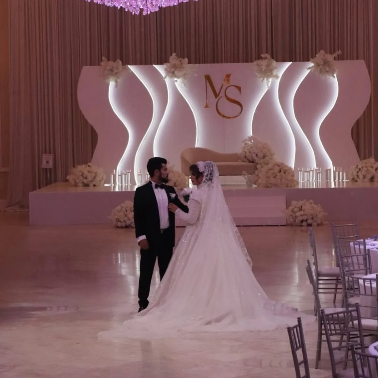 A bride and groom dancing in a decorated wedding reception hall with elegant lighting and floral arrangements.