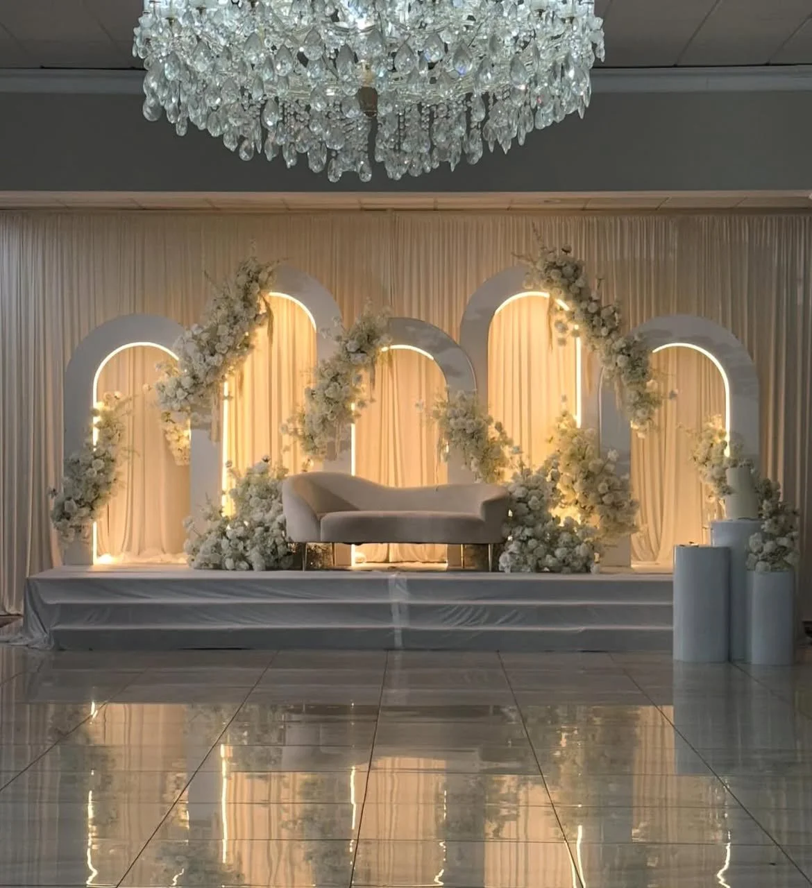 Elegant wedding or event stage decorated with white flowers, arch-shaped structures, a white chaise lounge, and a crystal chandelier overhead.